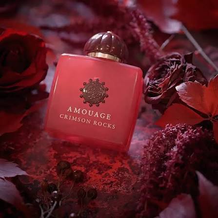 Amouage Crimson Rocks perfume bottle in a translucent coral-red color with a metallic gold emblem and lettering, surrounded by red roses and dried botanicals.