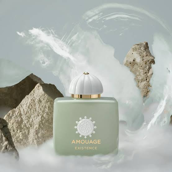 Amouage Existence perfume bottle in a pale sage green color with a white ribbed cap and gold lettering, set against a background of white waves and gray rocks.