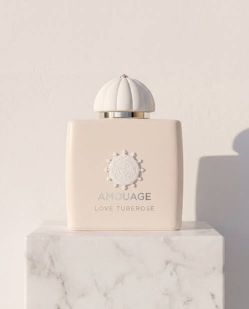 Amouage Love Tuberose perfume bottle in a pale cream color, resting on a white marble pedestal against a textured off-white wall.