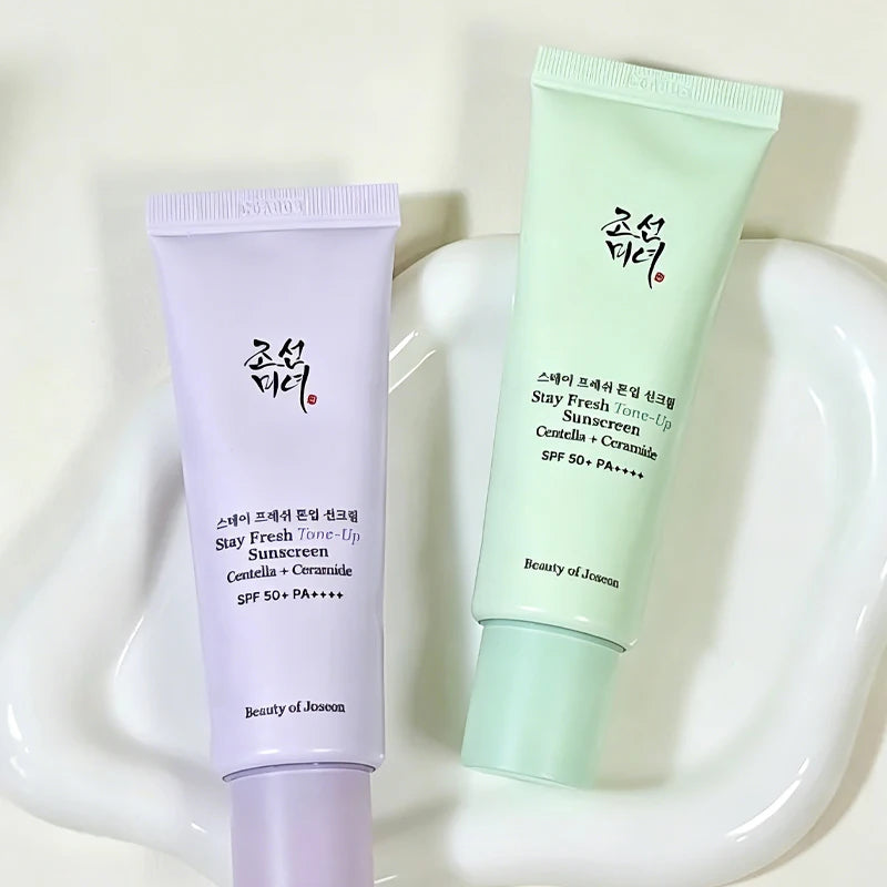 Two cosmetic sunscreen tubes from Beauty of Joseon displayed on a white ceramic dish. One tube is light lavender purple and the other is mint green, both with black and red Korean text and English text reading Stay Fresh Tone-Up Sunscreen SPF 50+ PA++++.