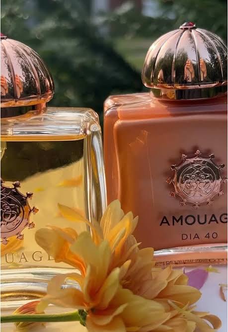 Two Amouage Dia 40 perfume bottles with metallic rose gold caps, one filled with golden-yellow liquid and the other with a peach-colored liquid, styled with a yellow flower in the foreground.