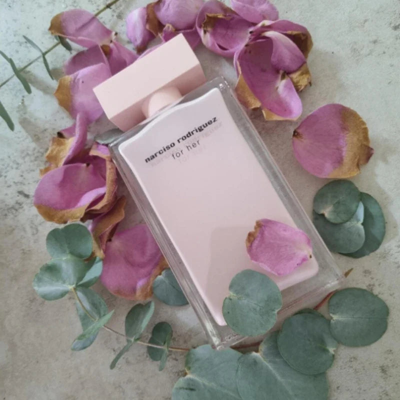 Narciso Rodriguez for Her perfume bottle surrounded by pink petals and green leaves on a textured surface