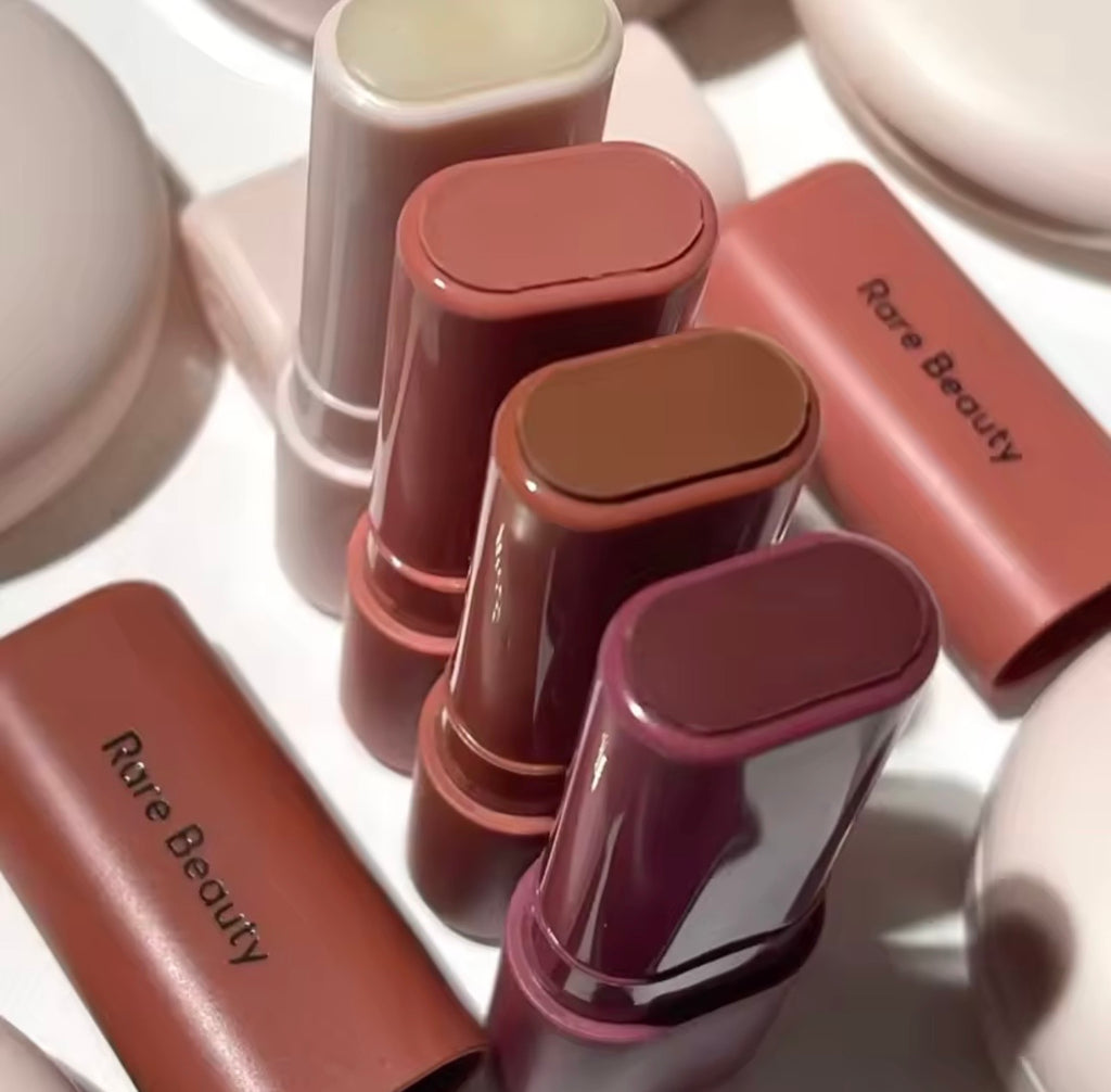 Four Rare Beauty lipstick bullets in warm neutral shades with their matching pink caps off, arranged on a white surface.