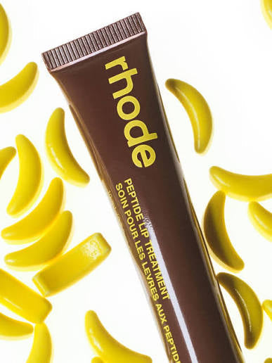 Rhode Peptide Lip Treatment in a brown squeeze tube with yellow lettering, surrounded by scattered yellow banana-shaped pieces on a white background.