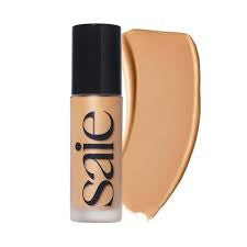 A bottle of SAIE liquid foundation with a black cap next to a large beige swatch of the foundation on a white background.