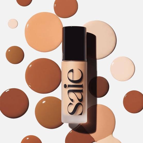 A bottle of Saie liquid foundation with a black cap and frosted glass, centered against a white background decorated with circular swatches of various skin-tone makeup shades.